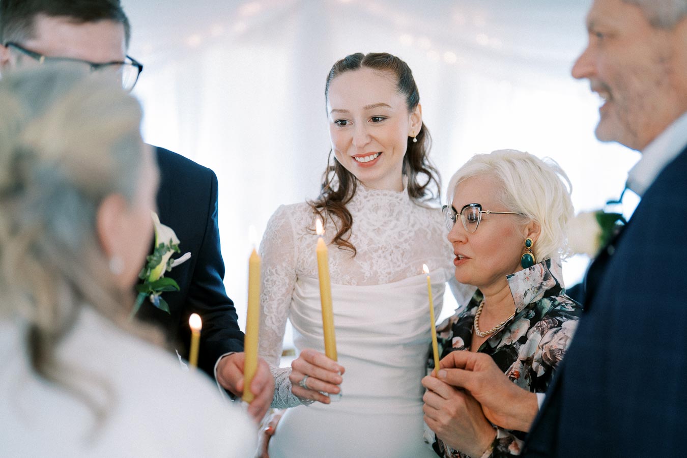 A bride in a white lace wedding dress stands with family members, each holding lit candles during a wedding ceremony.
