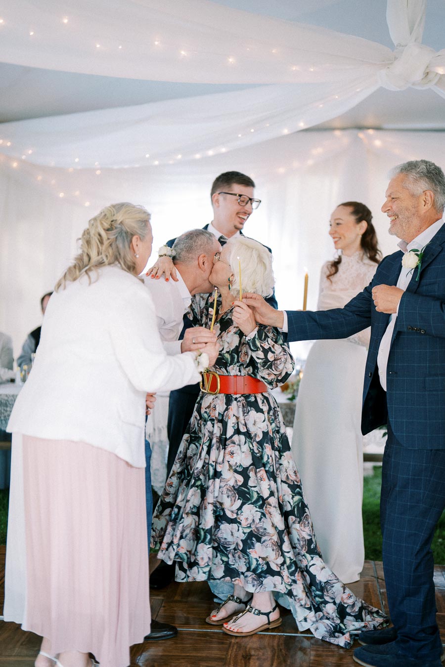A joyful group celebrating at a wedding reception under a white tent with twinkling lights. The scene captures a moment of closeness and happiness among the elegantly dressed guests.