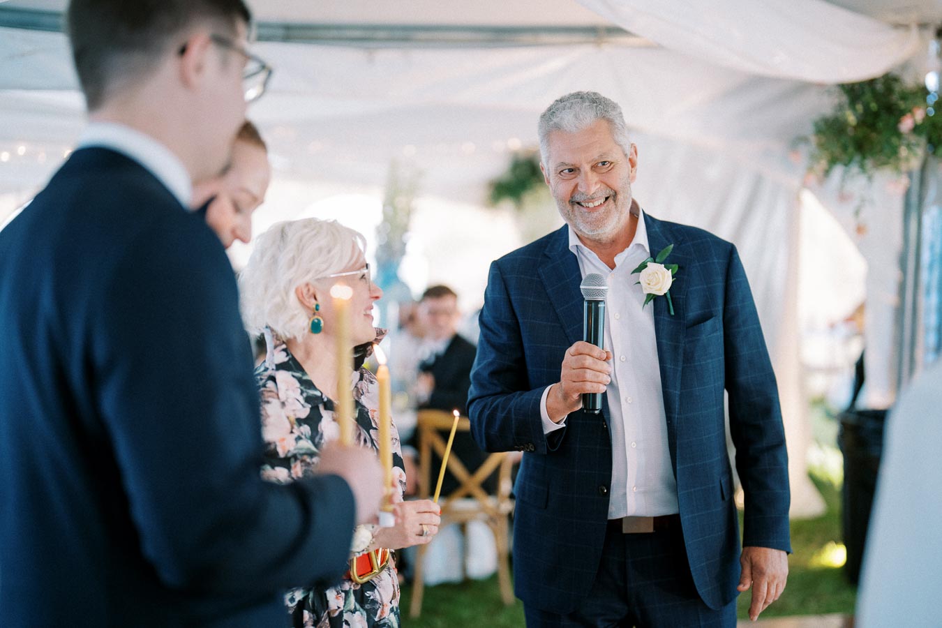Senior man in a blue suit holding a microphone and smiling at a wedding reception. Guests are gathered around and lighted candles can be seen in the foreground. The setting is a decorated tent with greenery and warm lighting.