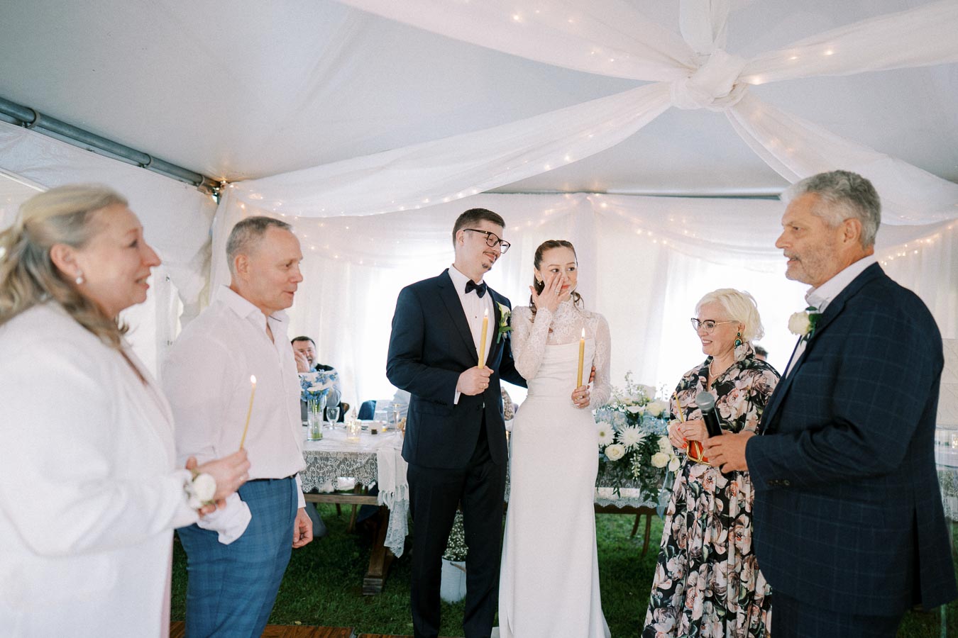A bride and groom surrounded by family and friends hold candles during a heartfelt wedding ceremony in an elegantly decorated tent.