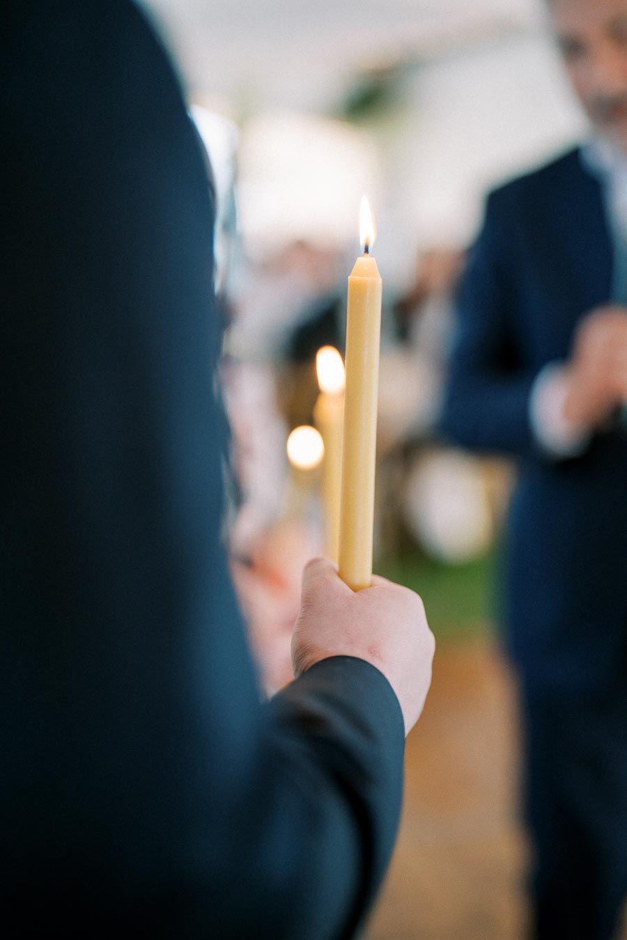 Person holding a lit candle at an event, with blurred figures in the background, symbolizing a ceremony or gathering ambiance.
