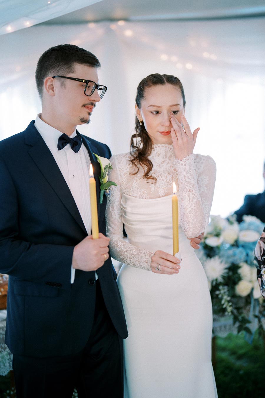 A bride and groom holding candles during a wedding ceremony, with the bride wiping away a tear. The scene is set in a softly lit venue with elegant floral arrangements in the background.
