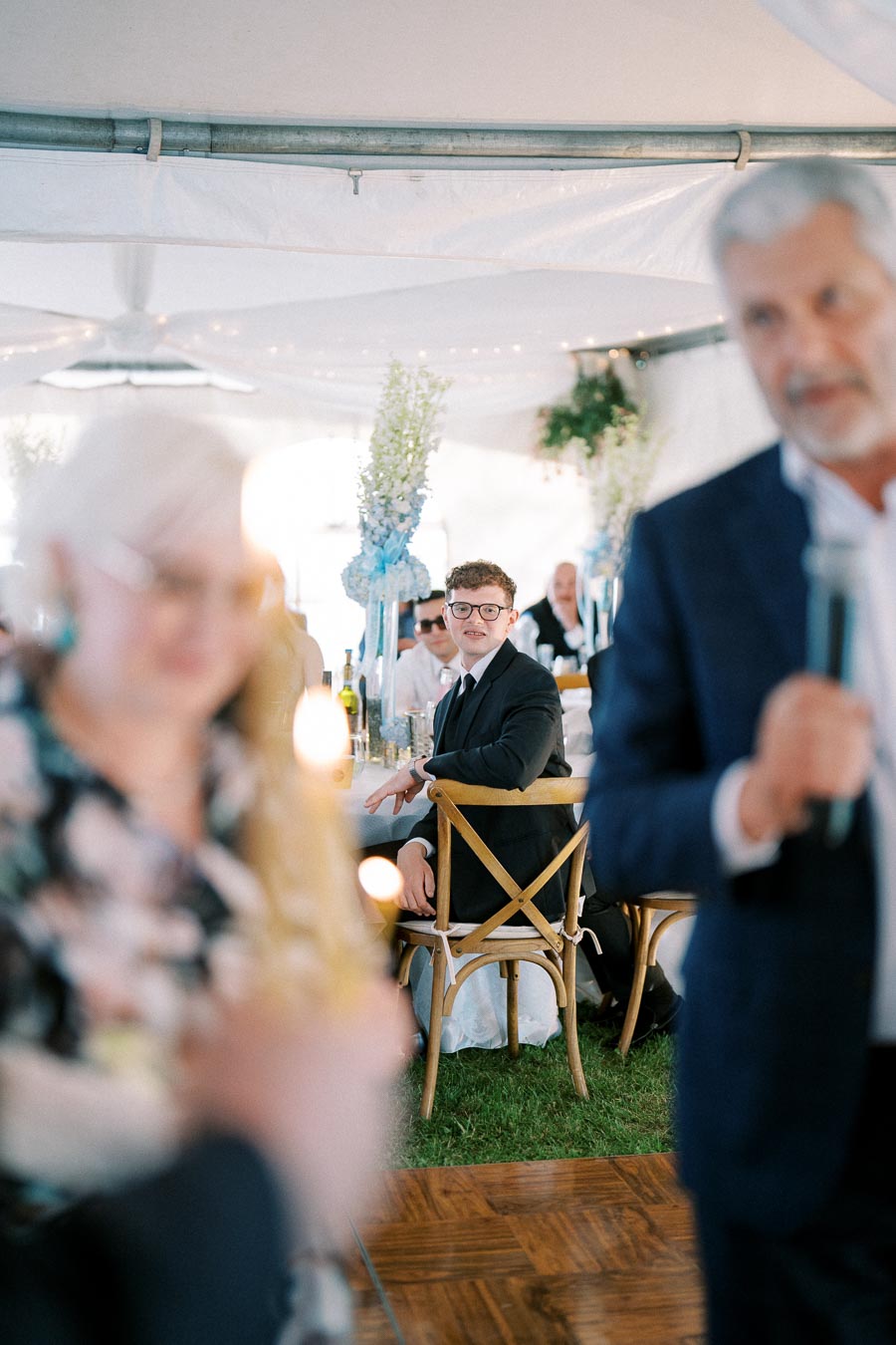 Wedding reception scene with a man in glasses seated at a decorated table, focusing on a speaker in the foreground. The atmosphere is elegant with floral centerpieces and ambient lighting under a tent.