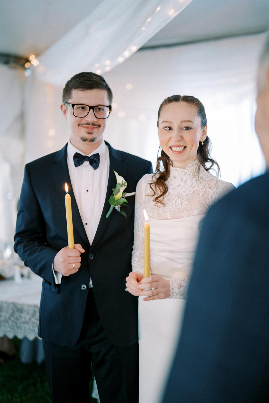 Couple holding candles at their wedding ceremony, dressed in formal attire with a lace wedding gown and suit, under a decorated tent.
