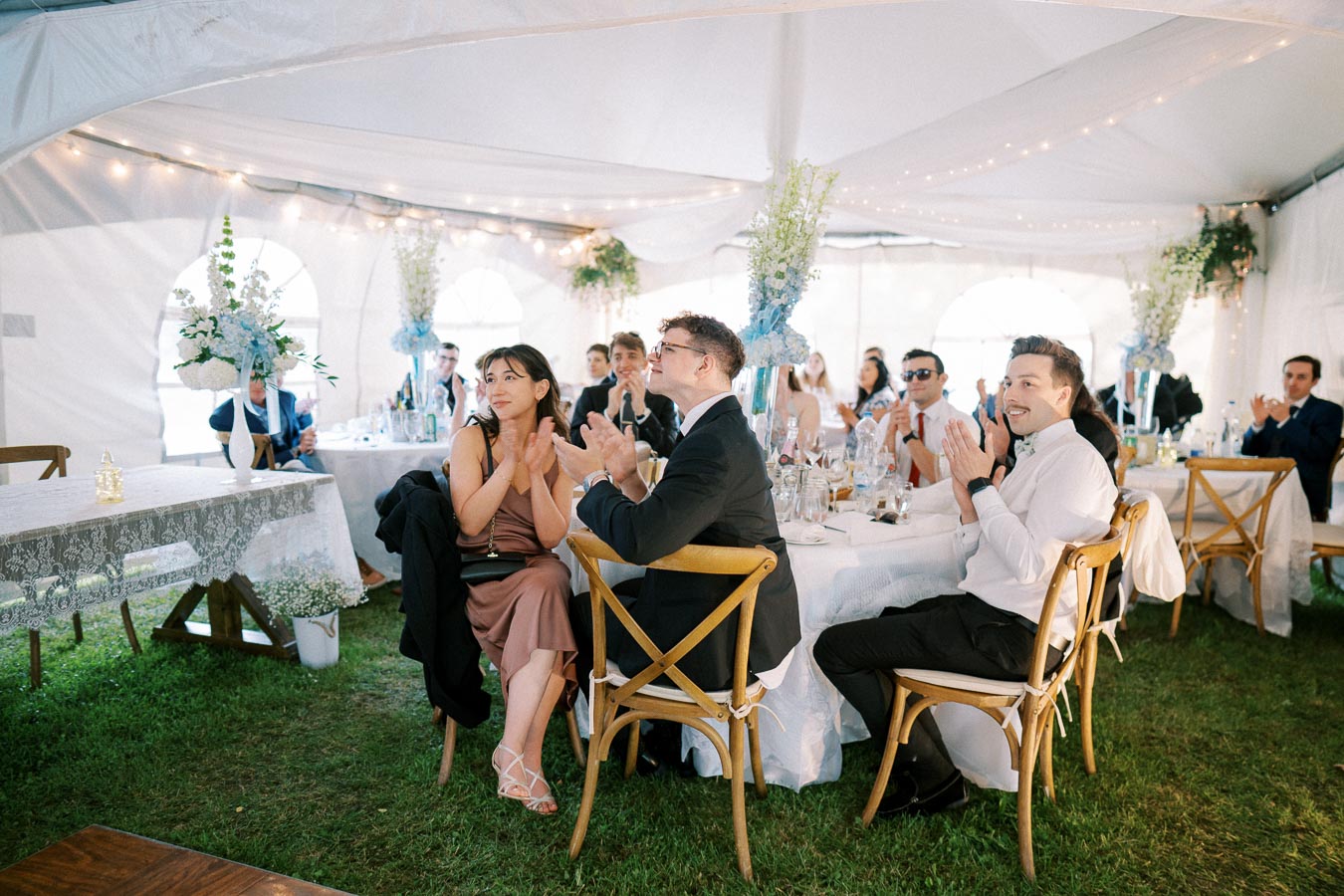 Guests clapping and smiling in a beautifully decorated wedding reception tent, featuring white tablecloths, floral centerpieces, and string lights.