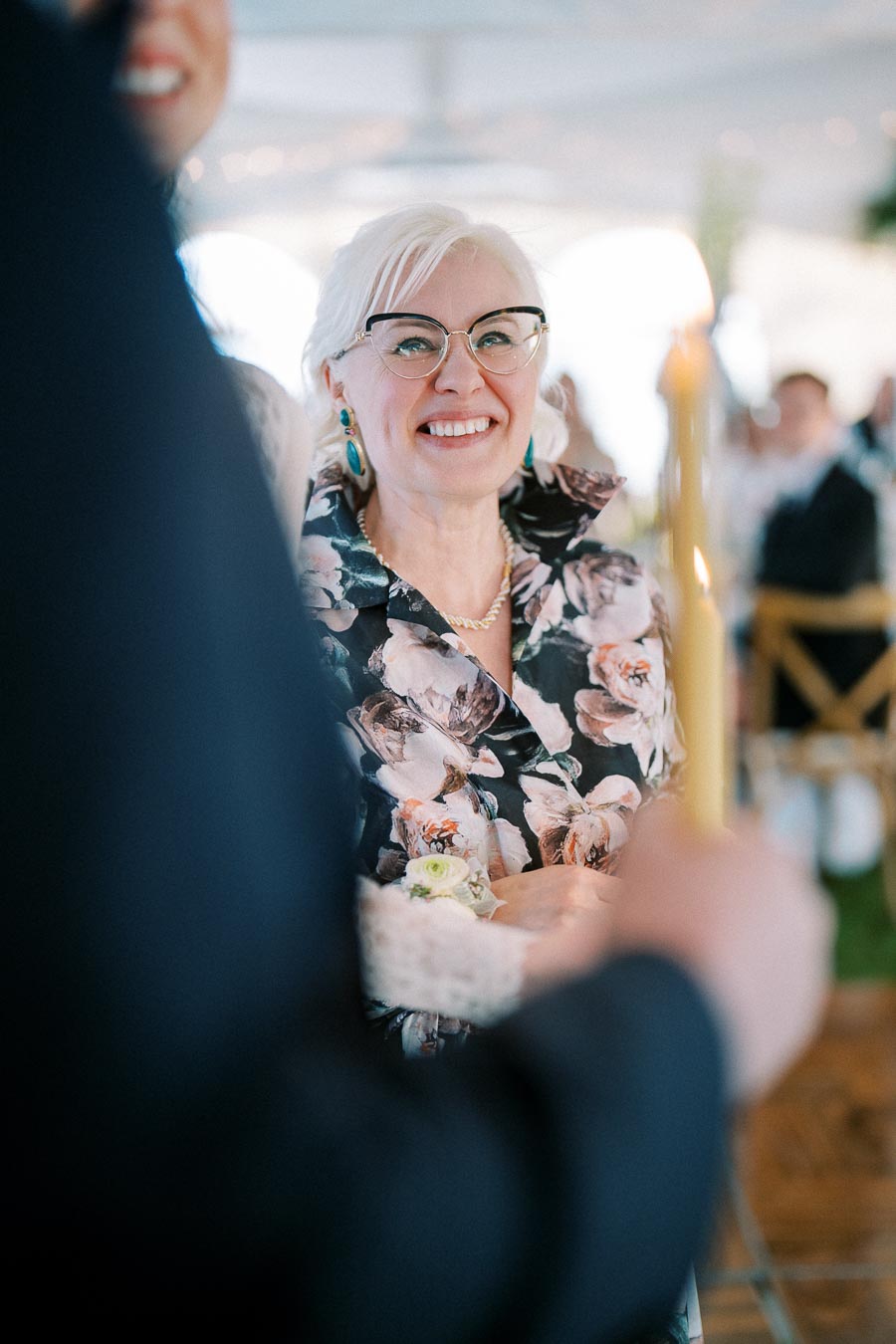 Senior woman with white hair and glasses smiling at an event, wearing floral attire and holding a candle.