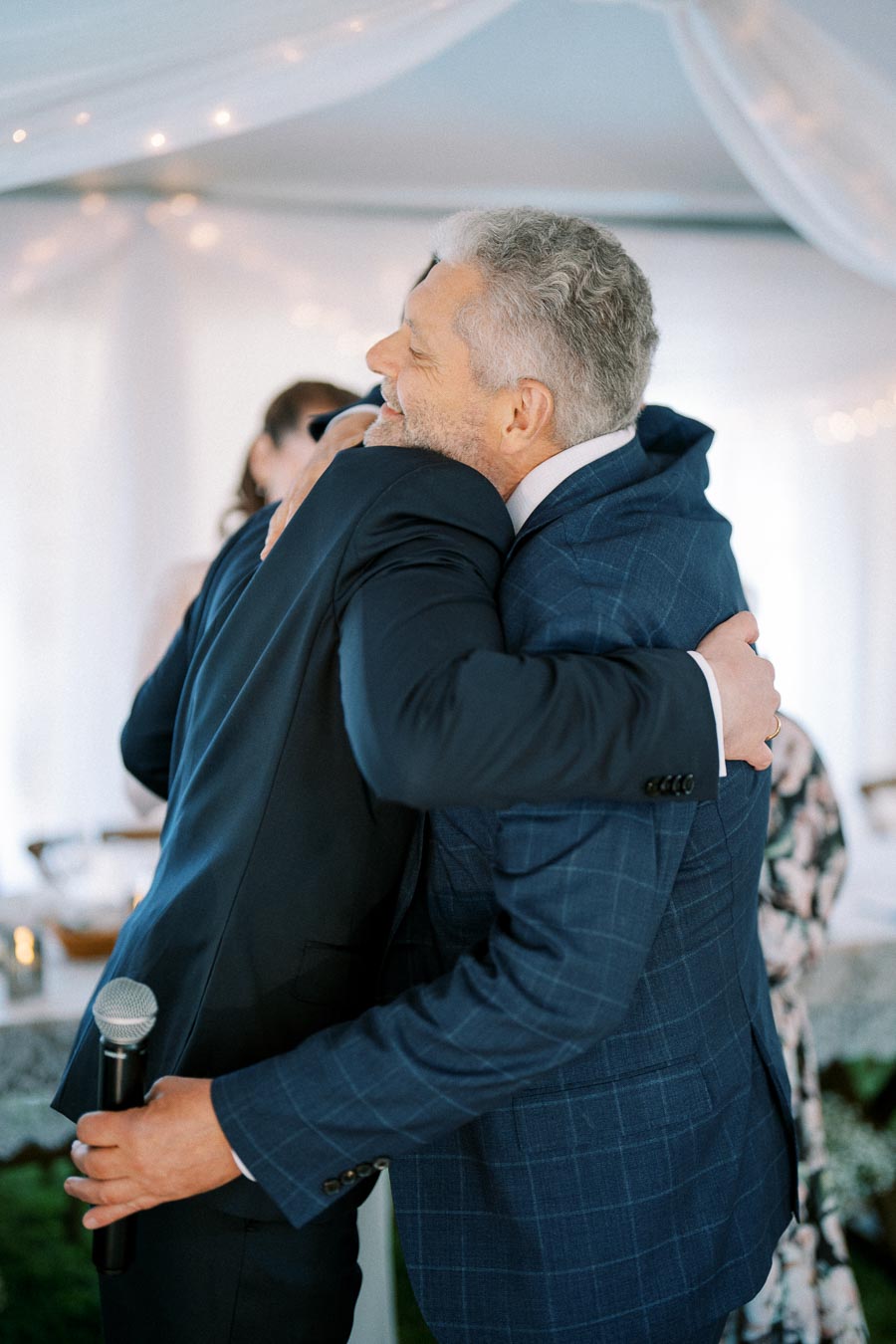 Two men in suits hugging at an indoor event, with one holding a microphone, under a draped ceiling with soft lighting.