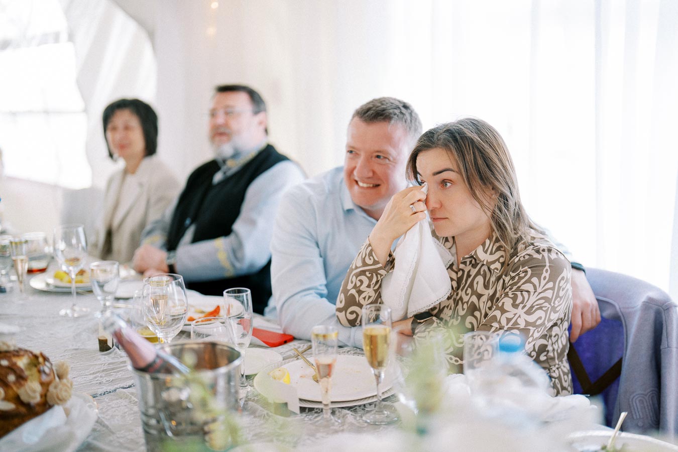 A group of people sitting at a dining table during a social event. One person, dabbing their eyes with a napkin, sits beside someone smiling warmly. The table is set with various dishes, drinks, and decorations, creating a festive atmosphere.