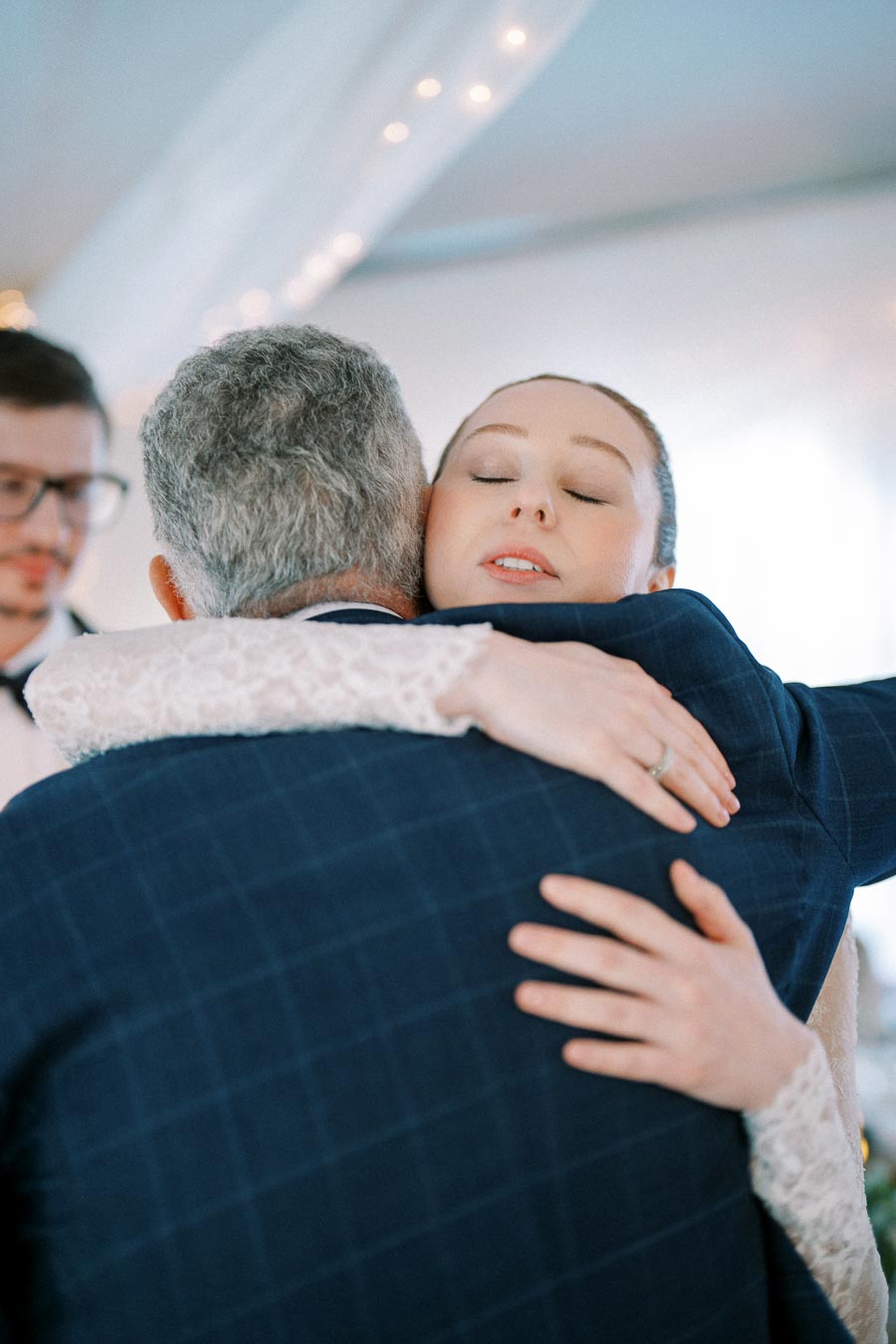 A bride in a white wedding dress embraces an older man in a dark suit during a wedding ceremony, with soft lighting and a blurred background.
