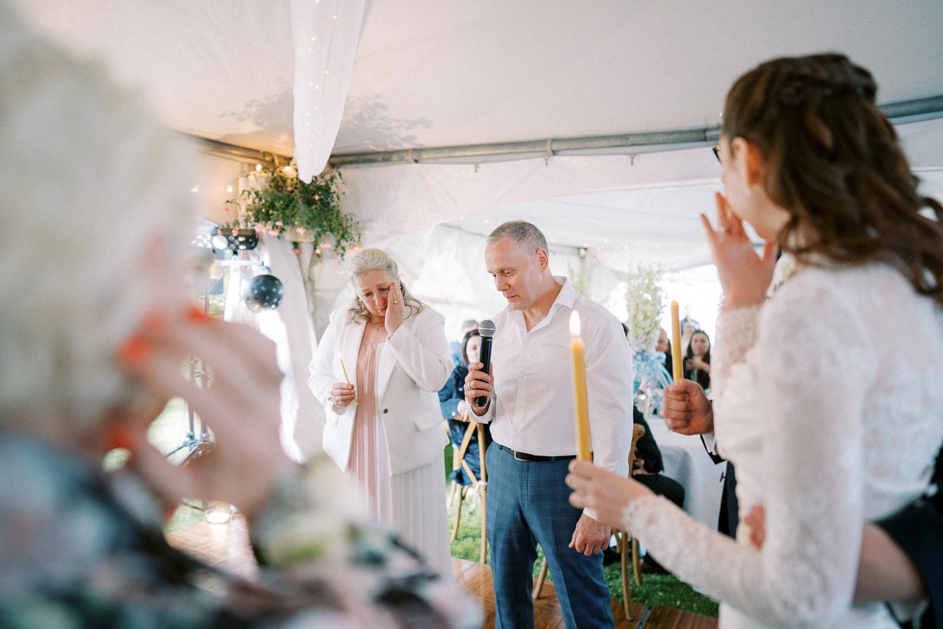 A heartfelt moment during a wedding ceremony under a tent, featuring a man speaking into a microphone and a woman wiping away tears, both holding candles, with guests watching in the background.