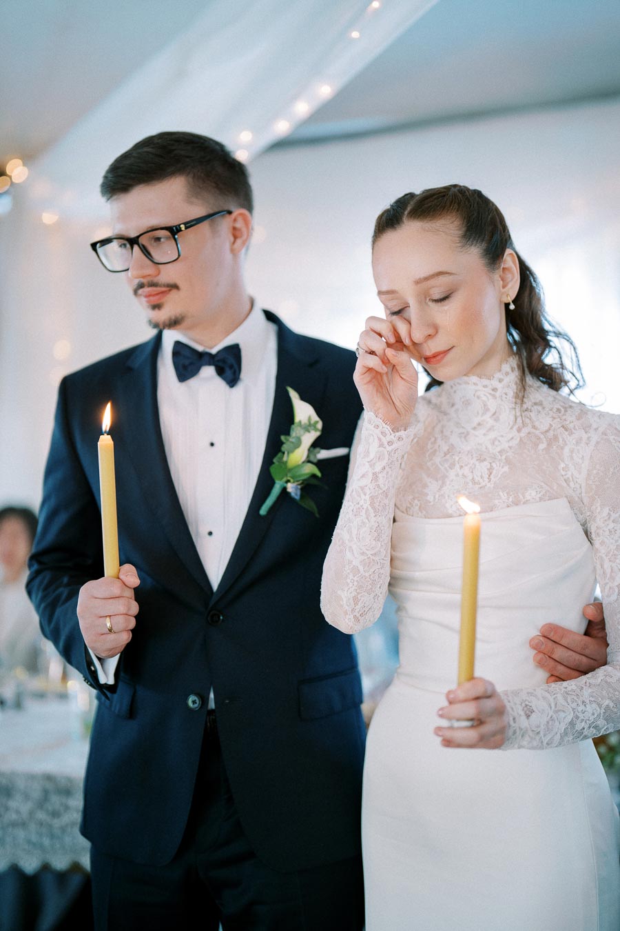 A bride and groom holding candles during a wedding ceremony, with the bride wiping away a tear while the groom stands supportively beside her. The groom is wearing a navy suit with a bow tie, and the bride is in a lace wedding gown.