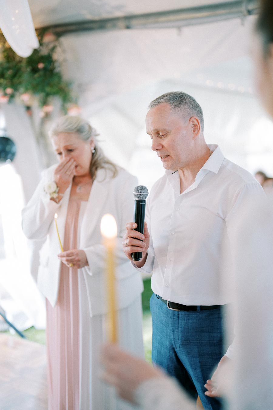 Elderly woman wiping tears and holding a candle, while a man in a white shirt speaks into a microphone during an emotional event inside a decorated tent.