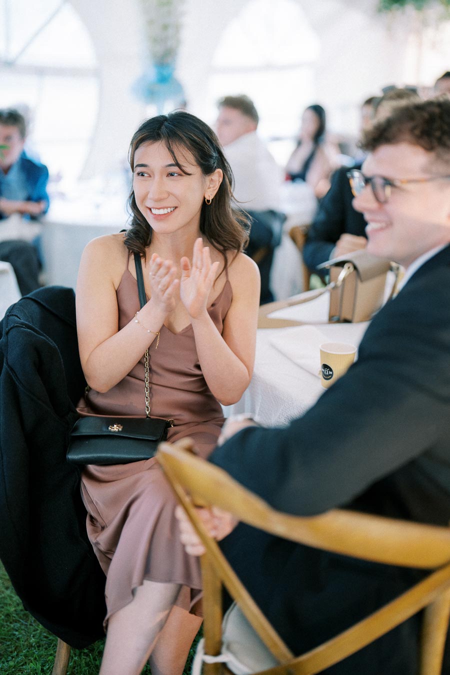 A young woman in a mauve dress smiles and applauds at a formal gathering, seated at a table with guests in the background.