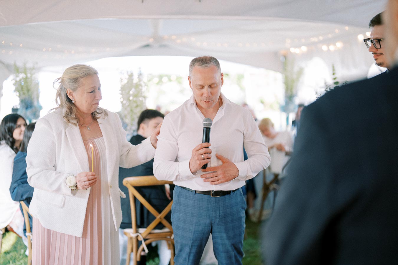 A man speaks into a microphone while a woman holds a candle, both participating in a ceremony under a white tent.