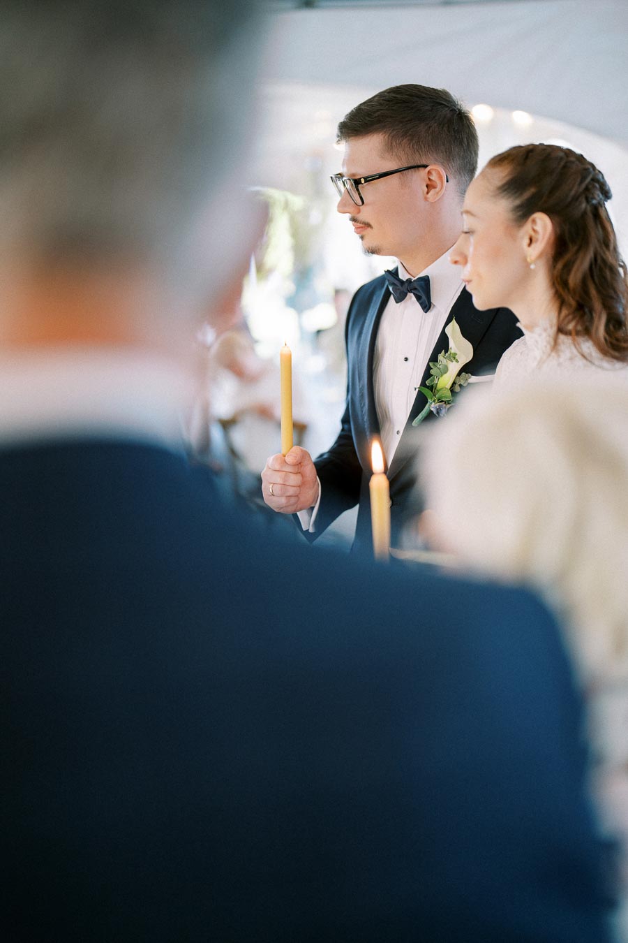 Bride and groom holding candles during a wedding ceremony, dressed in formal attire with floral decorations in the background.