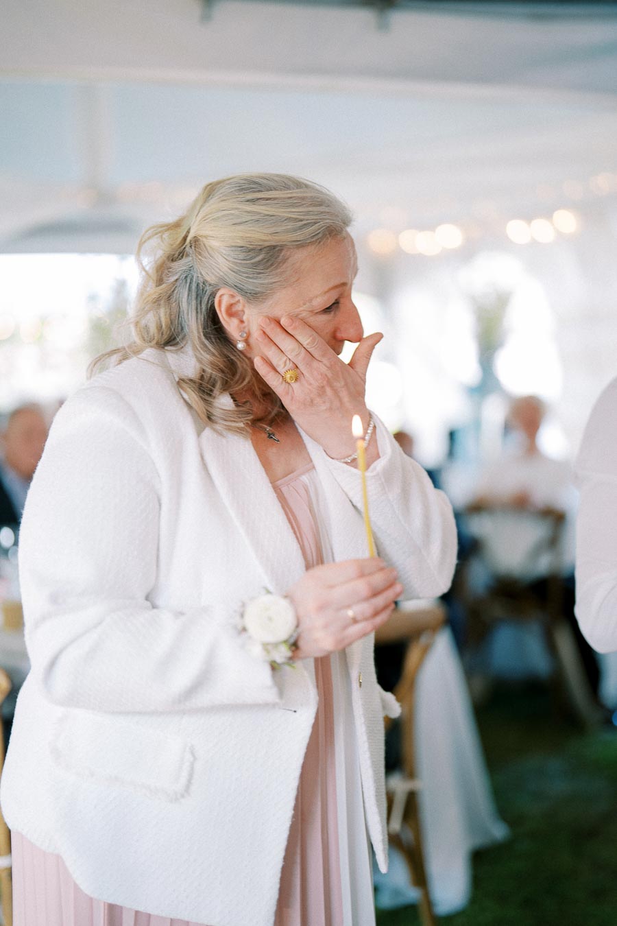 Elderly woman in a white jacket holding a lit candle, looking emotional at a formal event with blurred guests in the background.