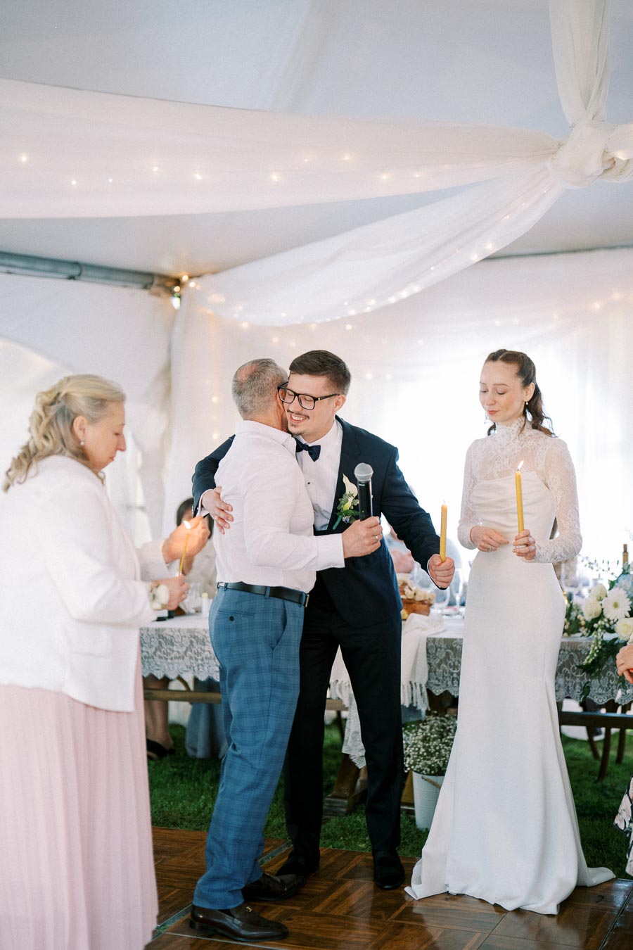 Wedding celebration scene with a groom in glasses hugging an older man while holding a microphone, and a bride in a lace wedding dress holding candles, surrounded by guests in an elegantly decorated venue with white drapes and string lights.
