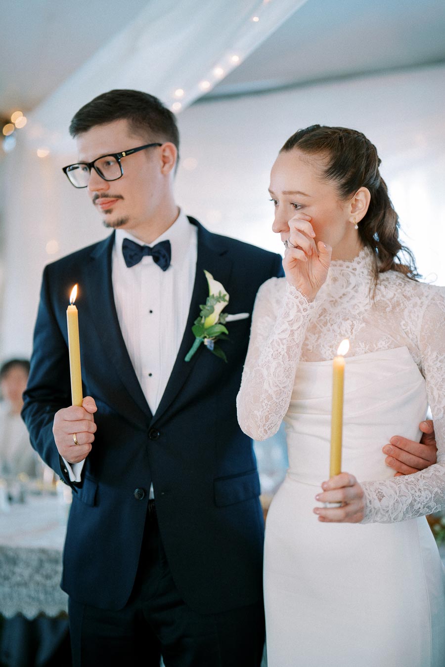 A bride and groom hold candles during an emotional wedding ceremony, with the bride wiping a tear from her eye. The groom is dressed in a formal suit and bow tie, and the bride wears a white lace gown.