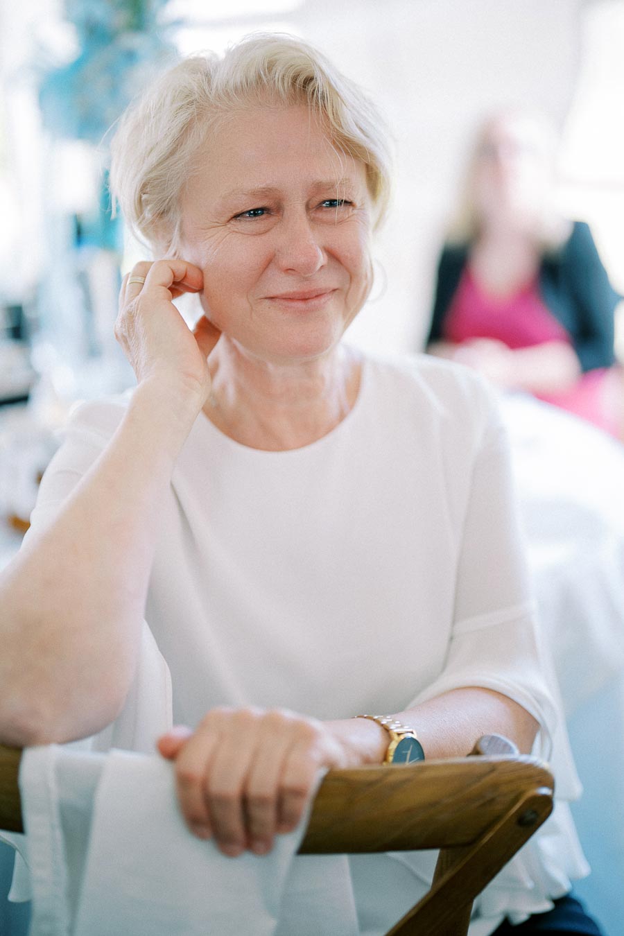 Elderly woman smiling while seated, wearing a white blouse and a watch, in a softly lit room with blurred background.