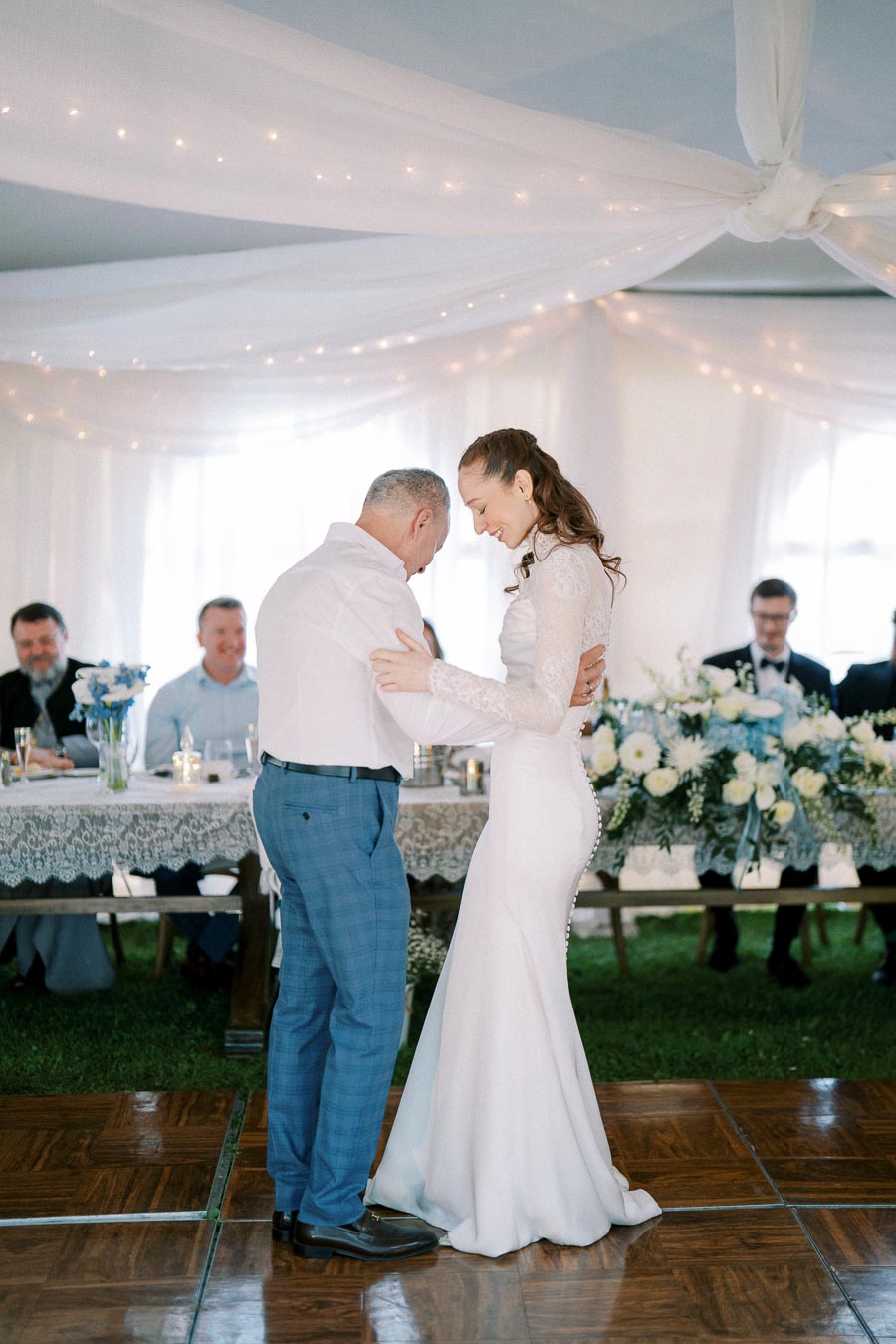 Father and bride sharing a dance at a wedding reception, surrounded by guests seated at a beautifully decorated table with floral arrangements.