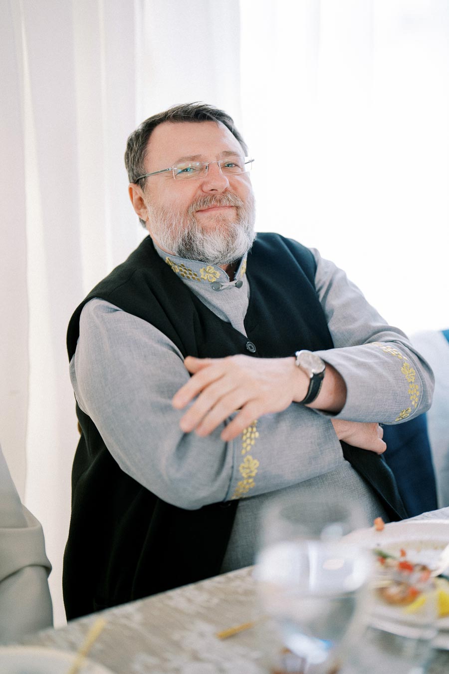 Man in traditional gray and black attire sitting at a table, smiling with arms crossed.