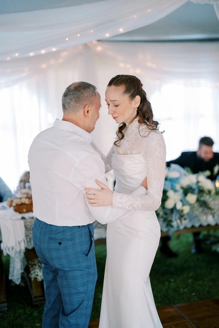 A bride in a white lace wedding dress shares a dance with an older man, both appearing emotional, inside a beautifully decorated venue with white drapes and string lights.