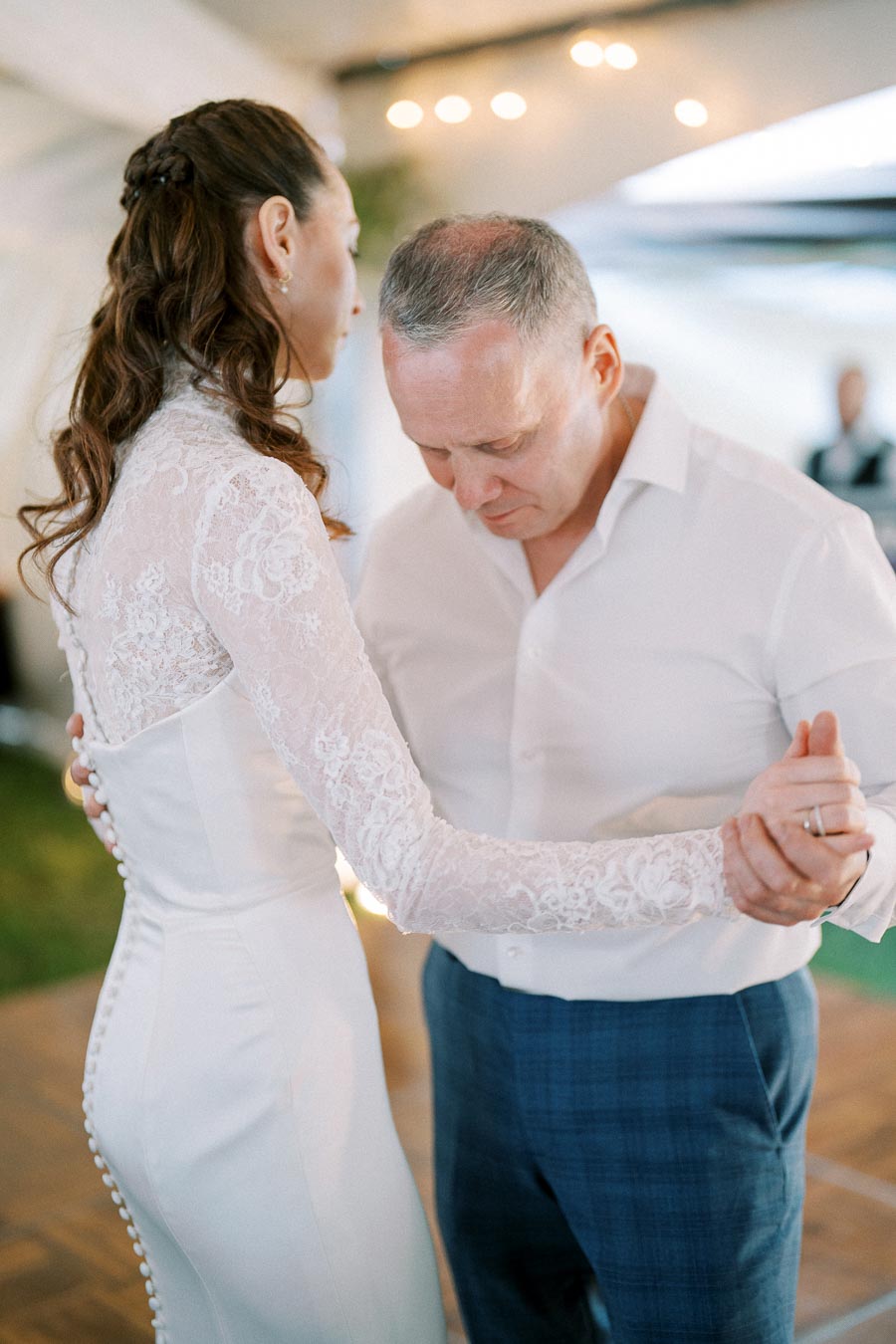 A couple shares a tender moment while dancing, with the woman wearing a lace wedding dress and the man in a white shirt and blue trousers, set in a softly lit event space.