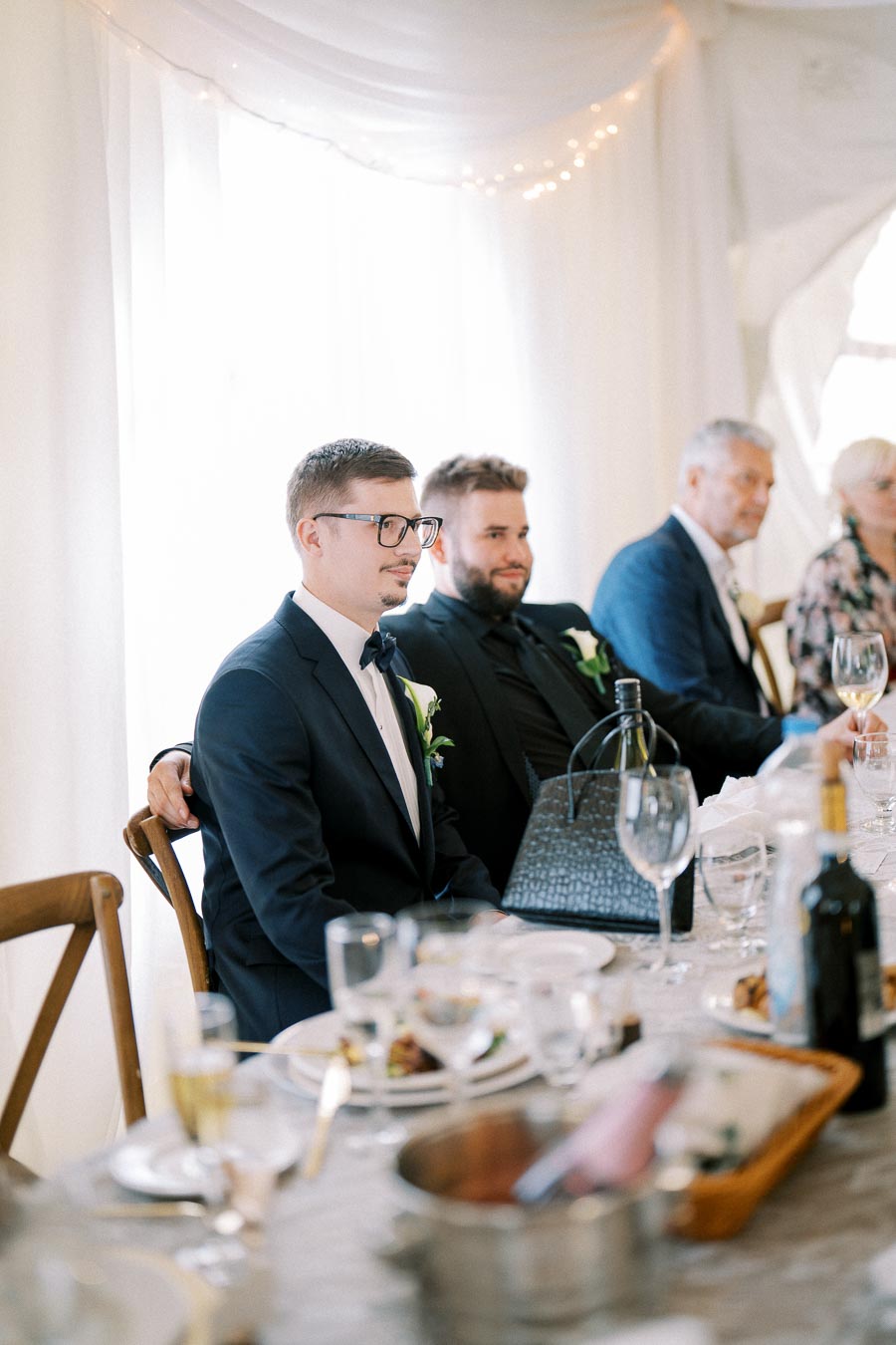 A group of well-dressed individuals sitting at a wedding reception table, showcasing elegant table settings, wine glasses, and decorative floral arrangements, creating a sophisticated and festive atmosphere.