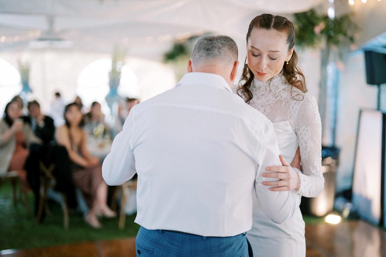 A bride in a lace wedding dress dances with an older man in a white shirt and blue pants at a wedding reception. Guests are seated in the background, attentively watching the dance.
