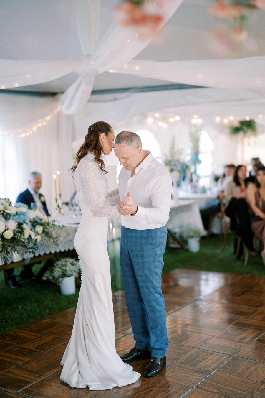 Couple sharing a romantic first dance at an elegant wedding reception, surrounded by beautifully decorated tables with floral arrangements and soft ambient lighting.