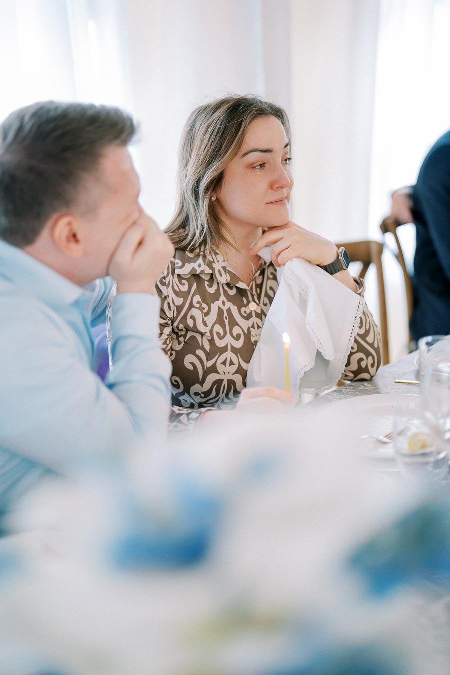 Two people sitting at a table in a contemplative mood during a formal event, one holding a lit candle and a napkin.