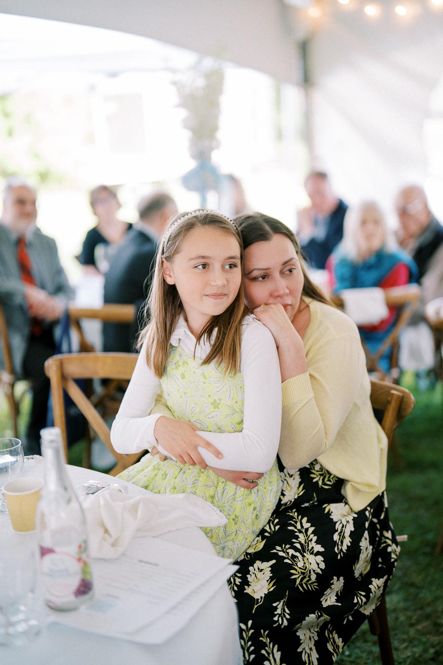 A woman and a girl seated at a table in an outdoor event, with the girl wearing a patterned green dress and the woman wearing a floral skirt, surrounded by other attendees in the background.