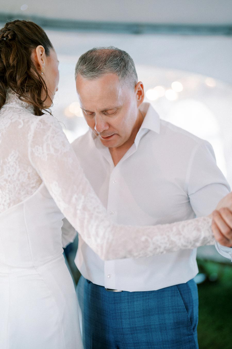 Elegant couple dancing at wedding reception, woman in lace dress and man in white shirt with blue pants, sharing an intimate moment.
