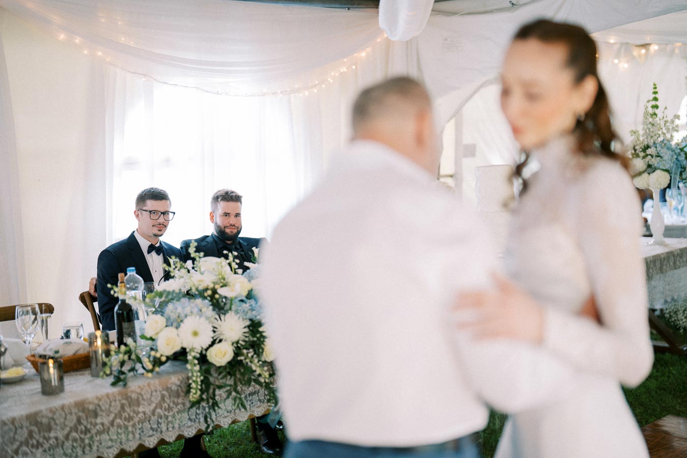 Wedding reception scene with a blurred couple dancing in the foreground. Two guests in formal attire sit at a decorated table adorned with white flowers and soft lighting, creating a festive and elegant atmosphere.