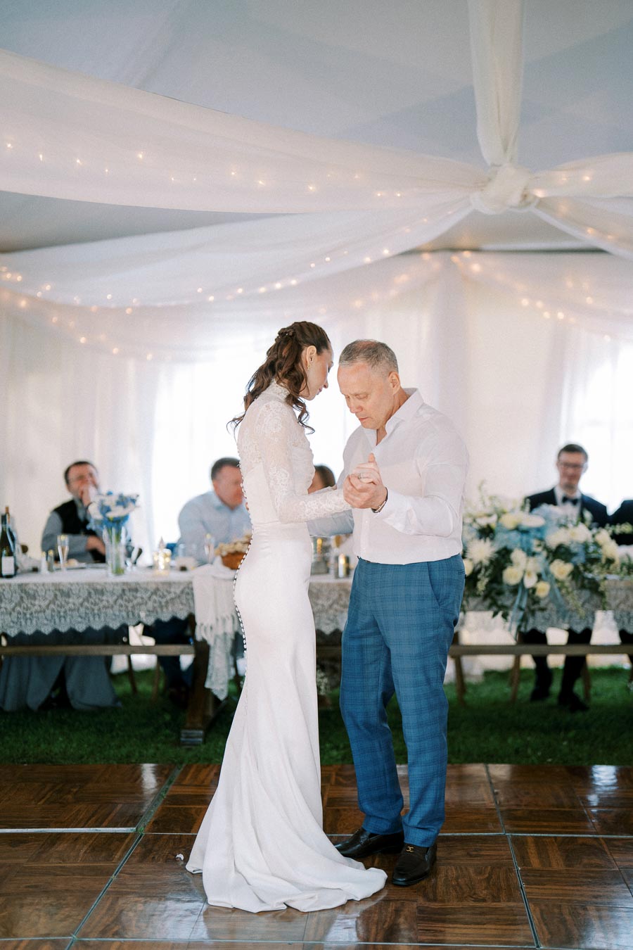 Father and bride share a dance at a wedding reception under a tent with white drapery and string lights, with guests seated at a decorated table in the background.