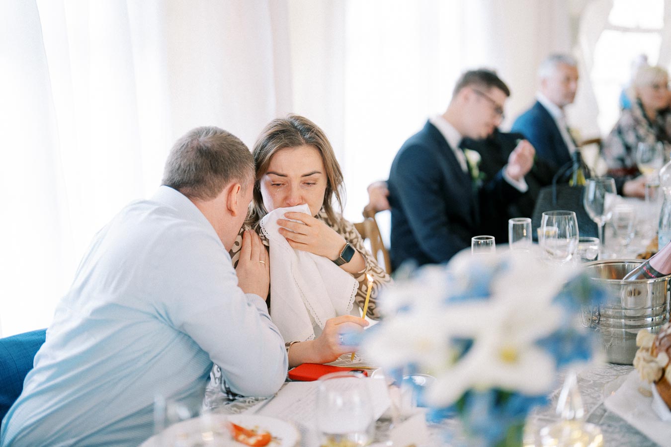 A group of people sitting at a decorated table during a formal event, with a woman wiping her face with a napkin as a man whispers to her, surrounded by glasses and floral arrangements, suggesting an emotional moment at a wedding or gathering.