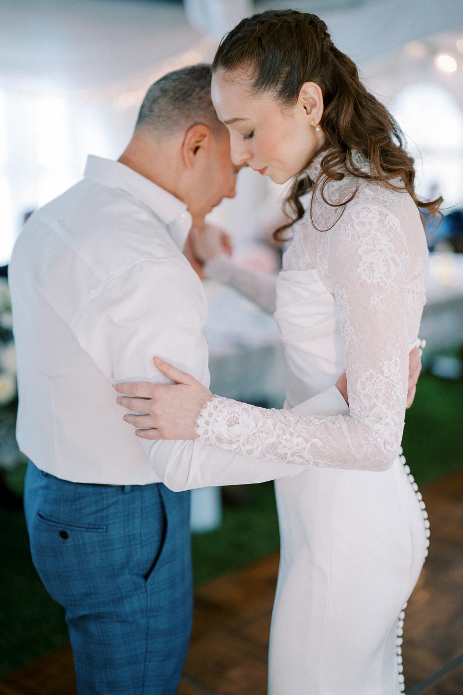 A bride in a lace wedding dress tenderly embraces a man in a white shirt and blue trousers during an intimate first dance at a wedding reception.