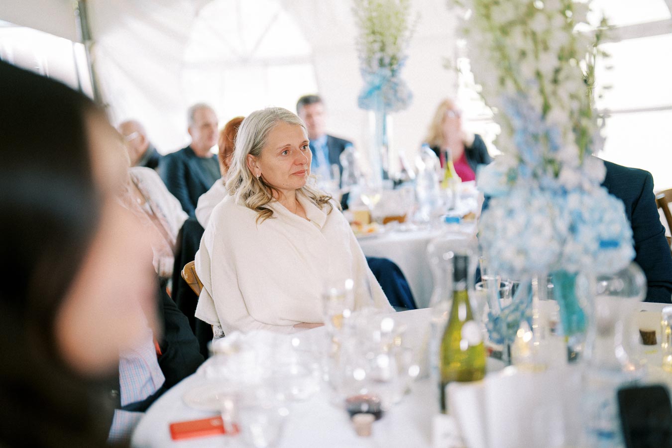 A woman seated at a table during a formal event, surrounded by elegant table settings and floral arrangements in a warmly lit venue.