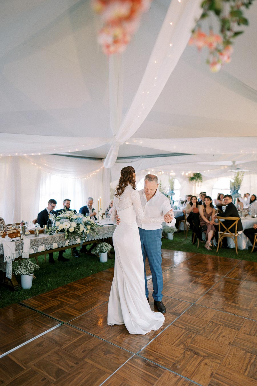 A bride in a white dress dances with her father under a decorated tent at a wedding reception, with guests seated at tables and flowers in the background.