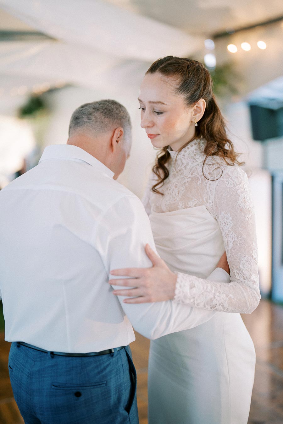 A bride in a lace wedding dress holds her partner during a romantic wedding dance, under soft lighting in a decorated venue.