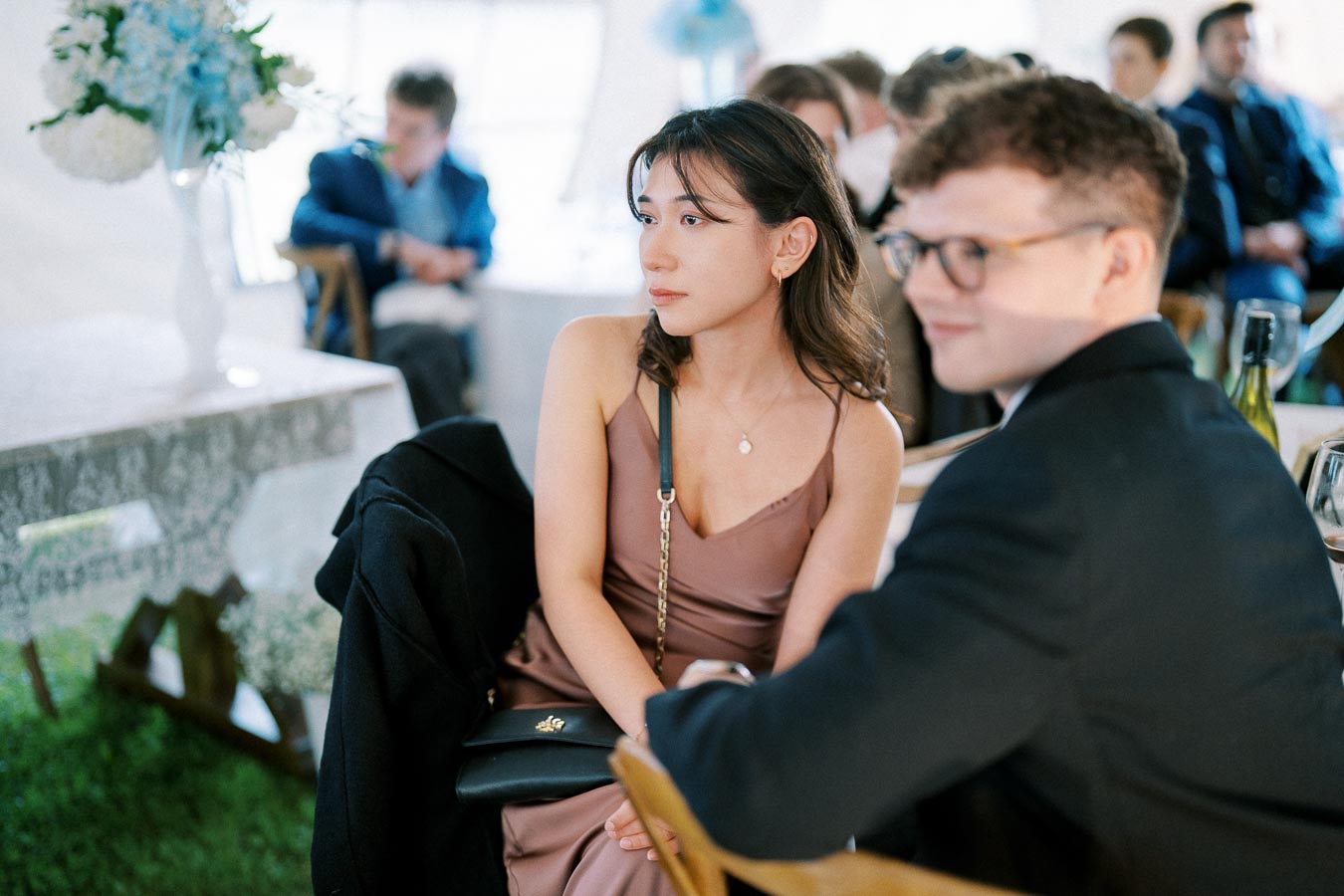 Elegant woman in a mauve dress attending a formal event, seated at a table with floral centerpieces and guests in the background.