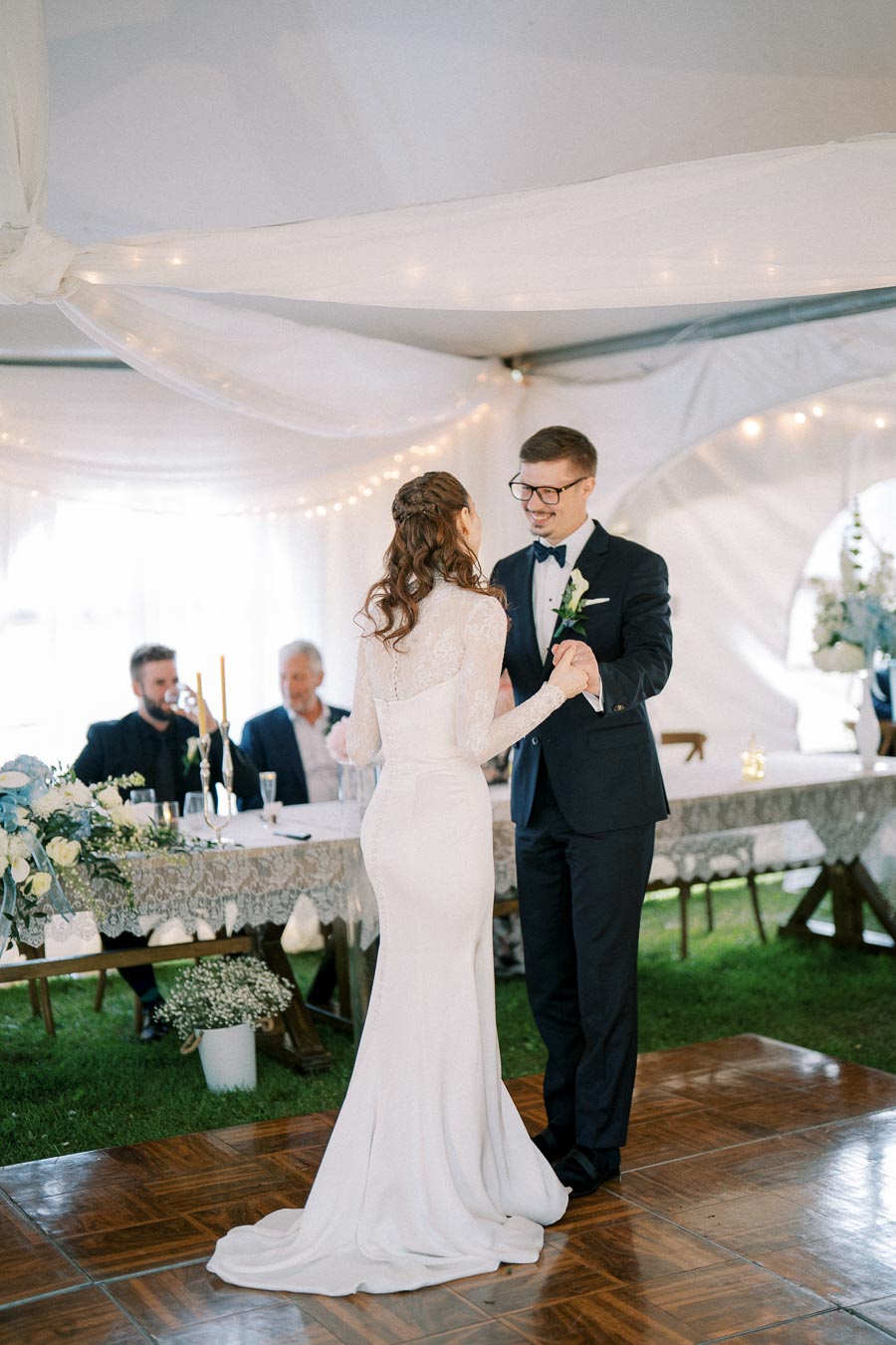 A bride and groom sharing their first dance at a wedding reception under a beautifully decorated tent, with guests seated at a table adorned with flowers and candles.