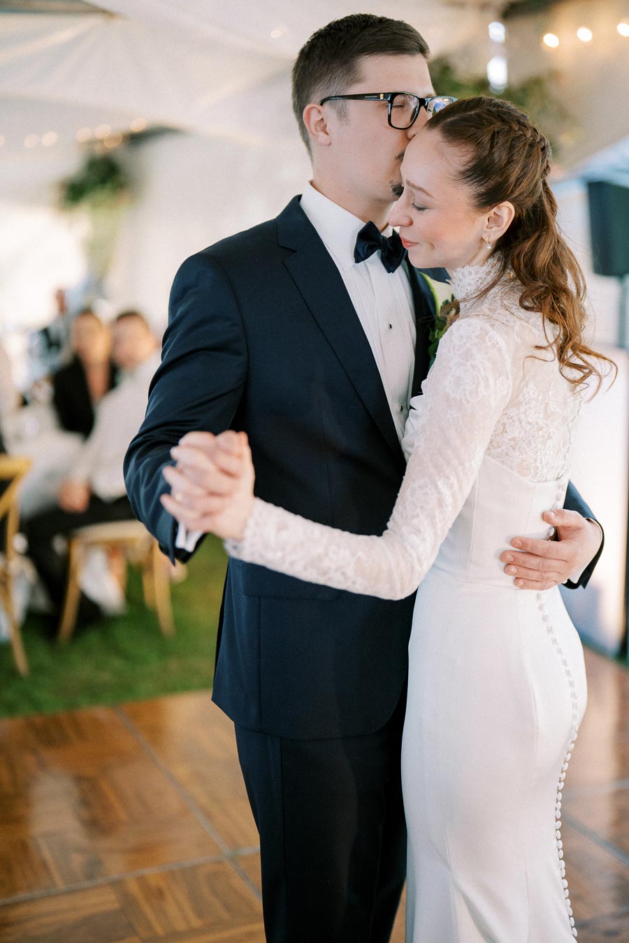 A bride and groom share a tender moment during their first dance at a wedding reception, with the groom in a navy suit and the bride in a white lace dress, creating a romantic and intimate atmosphere.