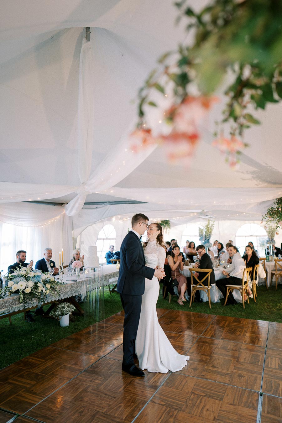 A couple shares their first dance at a wedding reception under a beautifully decorated tent, surrounded by seated guests and floral arrangements.