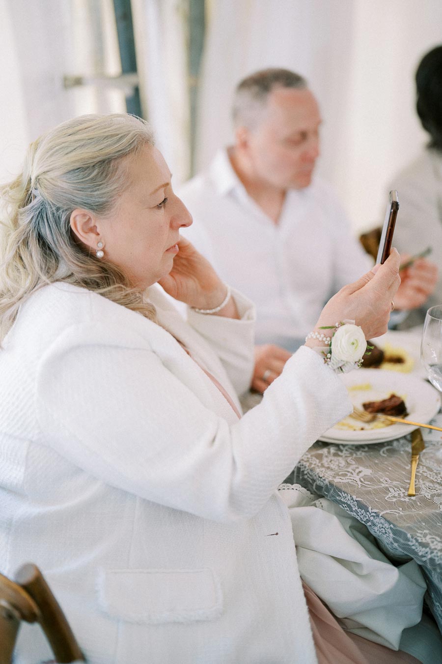 A woman in a white outfit using a smartphone at a formal dining setting, adorned with a floral bracelet.