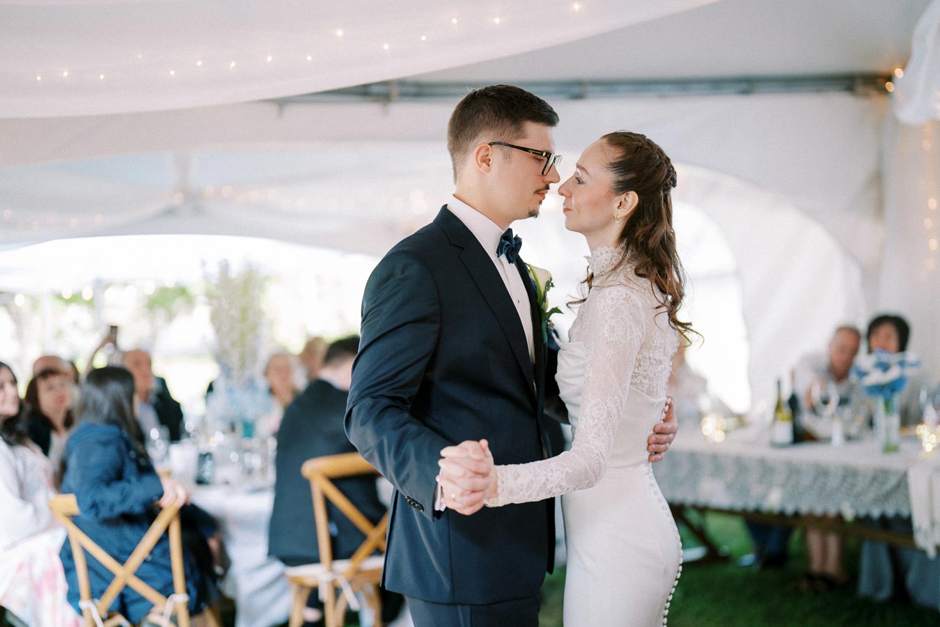 A bride and groom share a romantic first dance at their wedding reception under a white tent adorned with string lights, surrounded by seated guests.