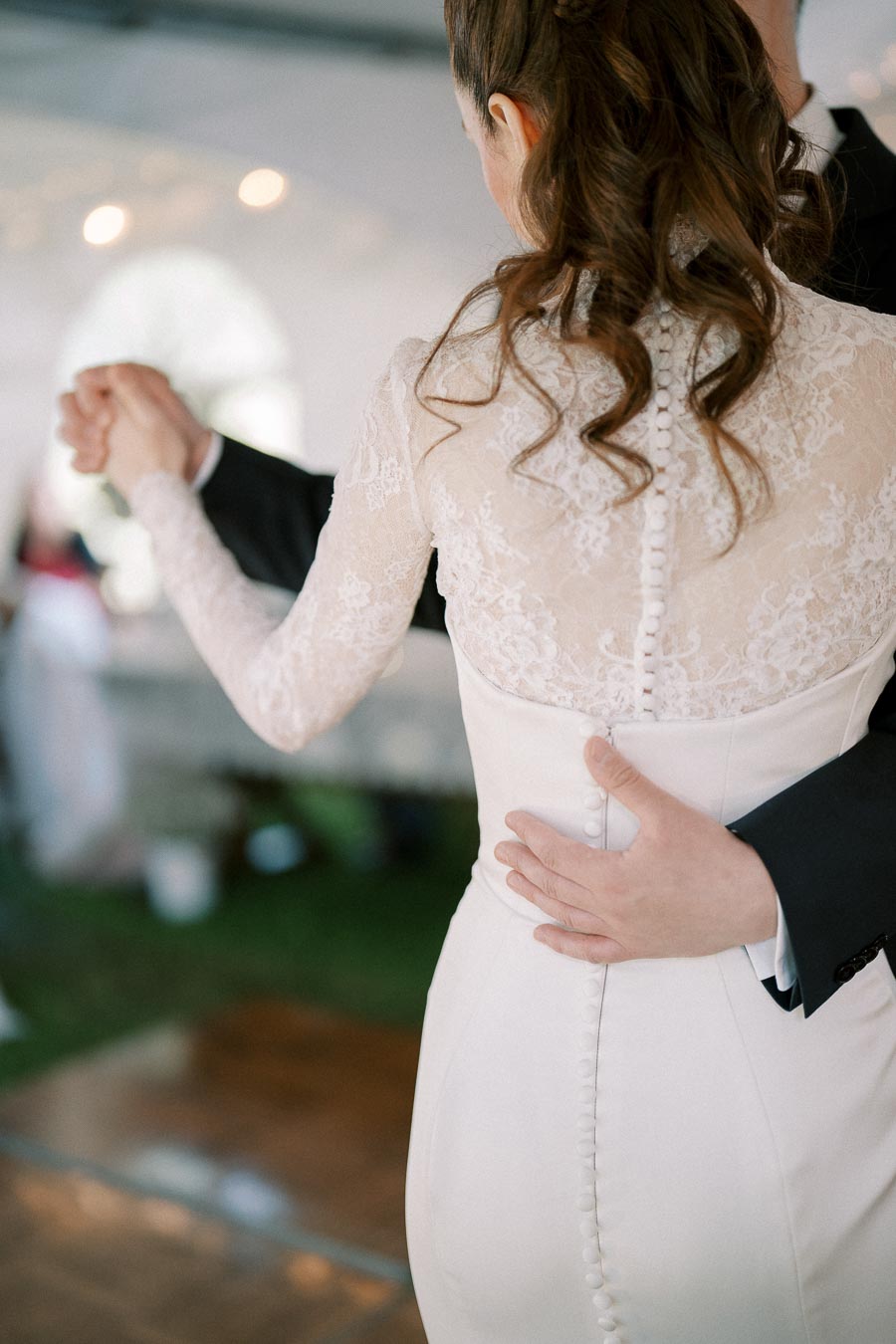 A bride and groom sharing a first dance at their wedding, with the bride wearing an elegant lace wedding dress featuring button details and the groom dressed in a classic black suit.