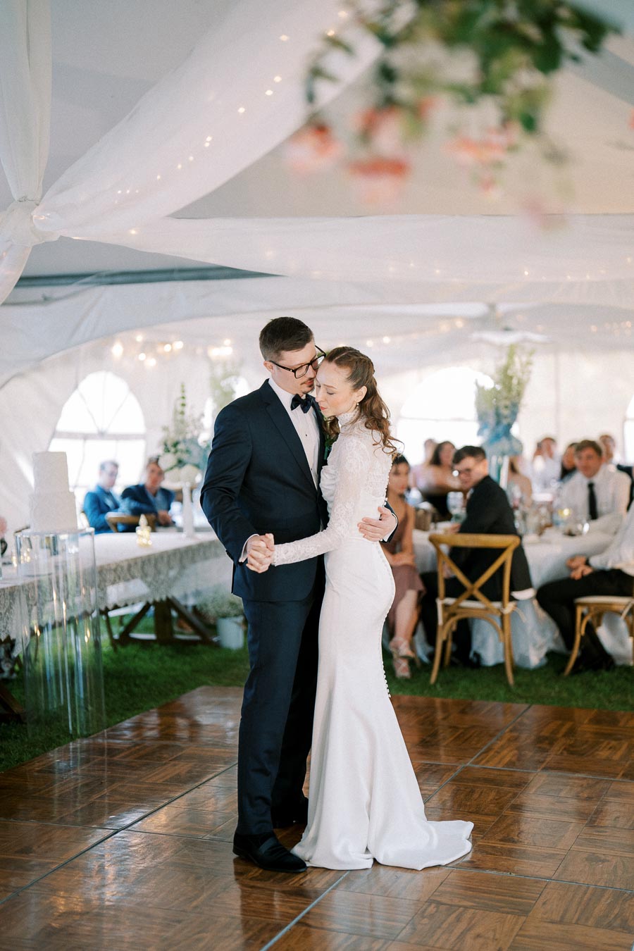 A bride and groom share their first dance at a wedding reception under a beautifully decorated tent, surrounded by guests seated at elegantly arranged tables.