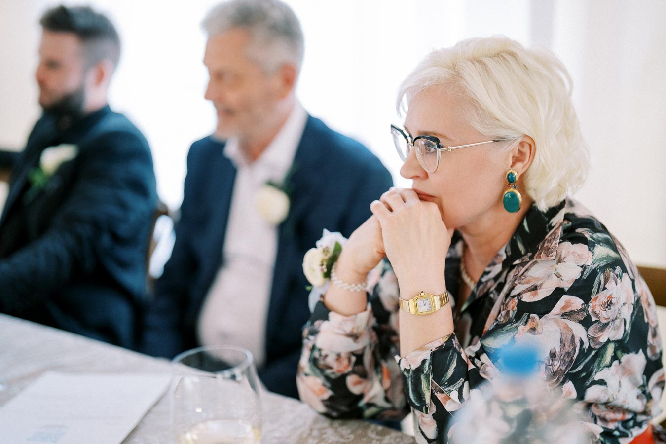 Elegant woman with short blonde hair and glasses, wearing a floral blazer and gold watch, sitting thoughtfully at a table with blurred figures in the background.