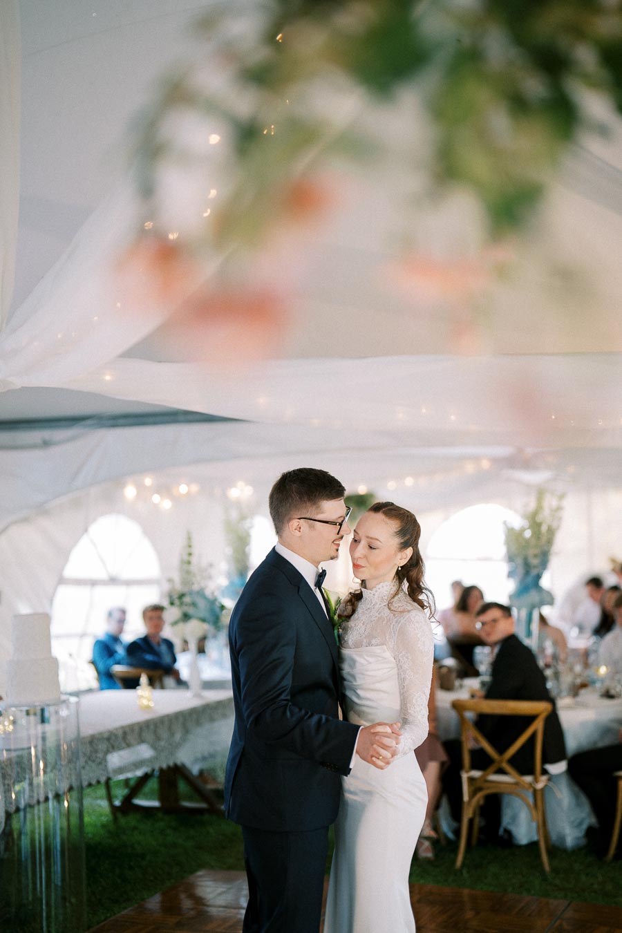 A couple sharing their first dance at a wedding reception inside a beautifully decorated tent, with guests seated at tables in the background.